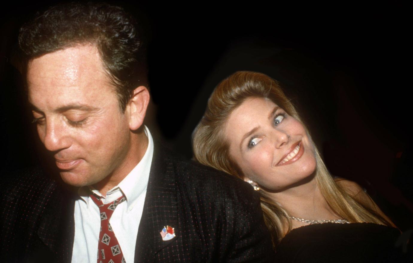 A smiling Christie Brinkley in a black dress, leans back on Billy Joel, who wears a black jacket, white shirt, and patterned tie.