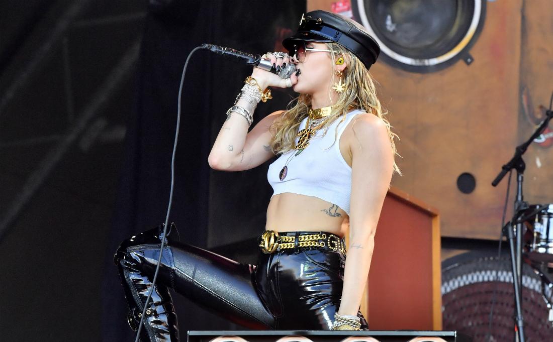 Miley Cyrus performs on stage in leather pants, cropped top, and black hat.