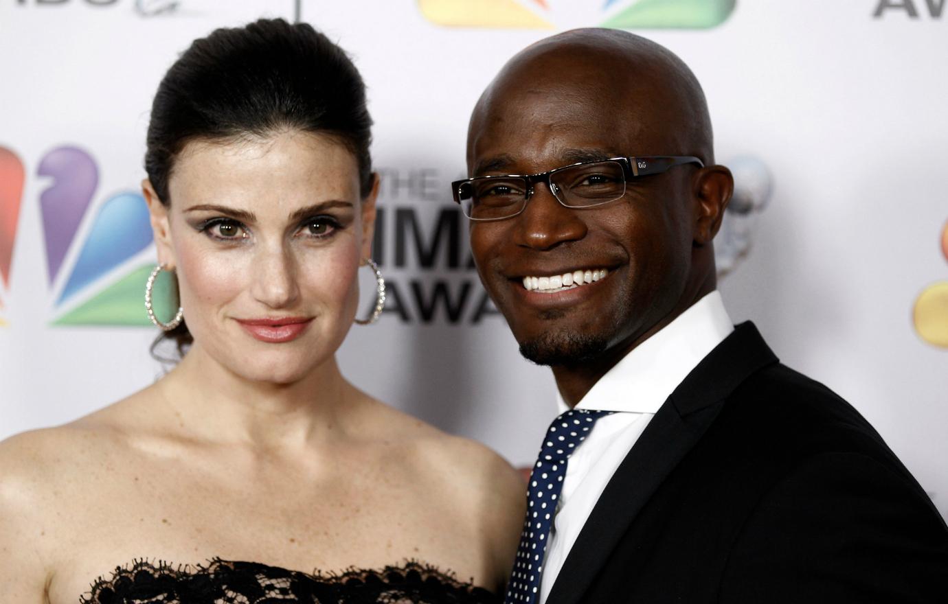 Idina Menzel, in a black dress, stands on the red carpet alongside Taye Diggs who wears a black jacet, white shirt, and polka dot black and white tie.