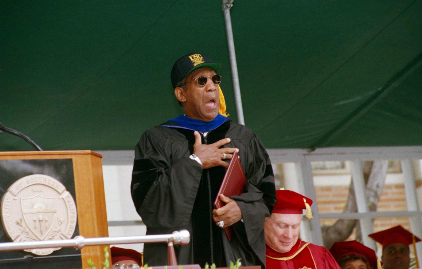 Bill Cosby stands on a stage in a black top.