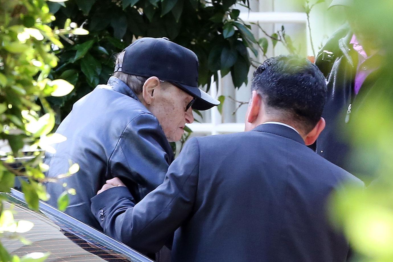 Larry King Wearing Baseball Cap Being Helped Out Of Car