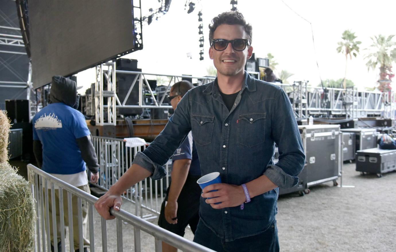 Evan Felker , in dark glasses, blue shirt, and holding a blue cup and smiles for the camera backstage at a concert.