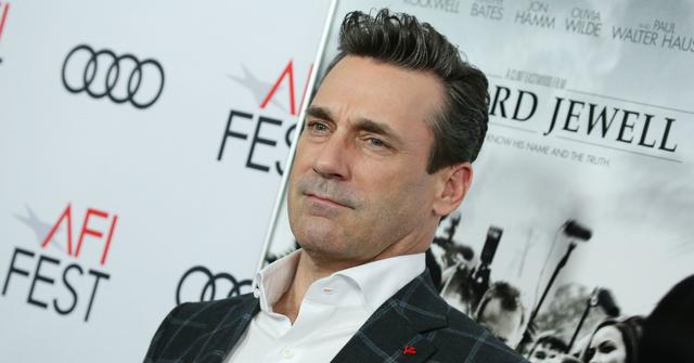 Jon Hamm, in a white shirt and check jacket, stands on the red carpet.
