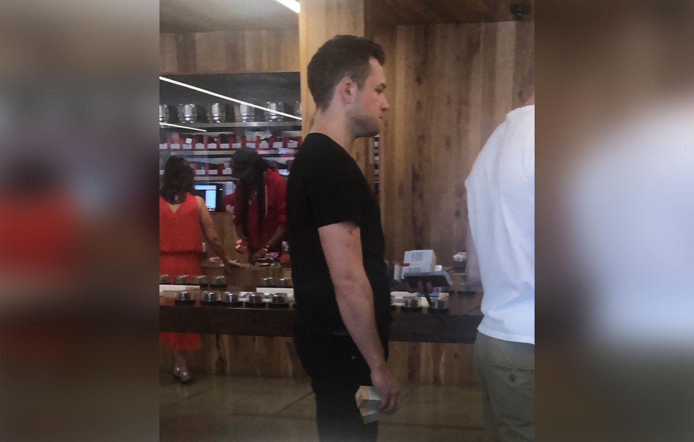 Taron Egerton Wearing Black T-Shirt and Black Jeans Standing In Line Inside a Medical Marijuana Store