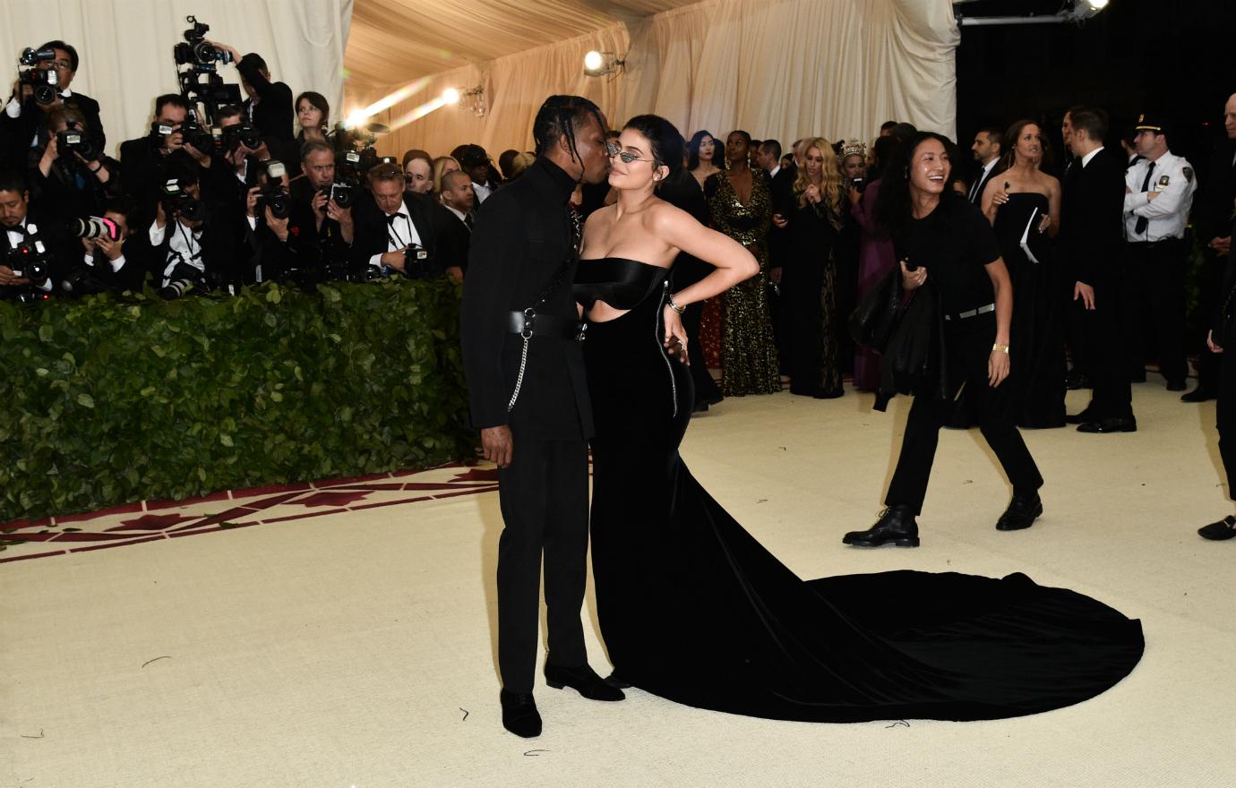 Travis Scott, in a black outfit, kisses Kylie Jenner on the cheek. Jenner wears a black dress and glasses.