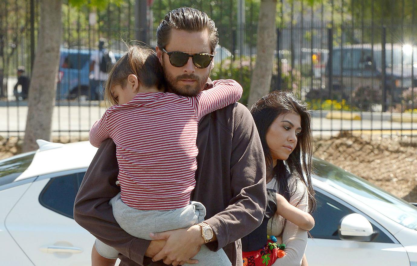 Scott Disick, in layered top and blue jeans, holds his child while Kourtney Kardashian, in blue jeans, walks behind him with their other child.