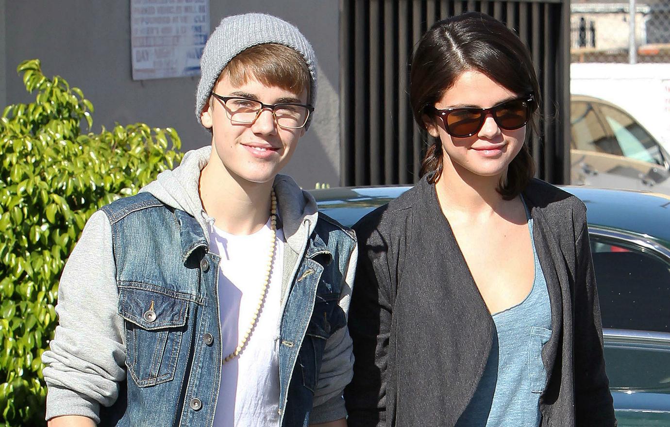 Justin Bieber, in glasses, beanie and jean jacket holds hands with Selena Gomex who wears a grey jacket, light blue top and blue jeans.