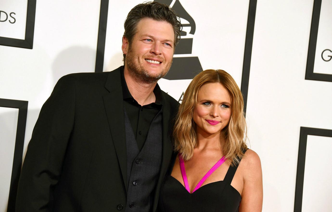 Blake Shelton, in a black suit, poses on the red carpet with Miranda Lambert who wears a black dress with pink trim.