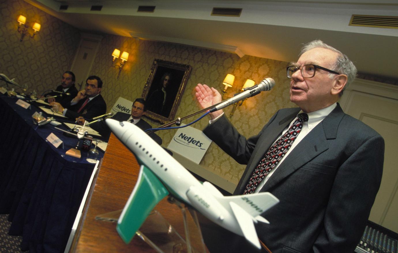 Warren Buffet, in a suit and tie, stands behind a podium giving a speech into a microphone. A small model airplane sits in the foreground.