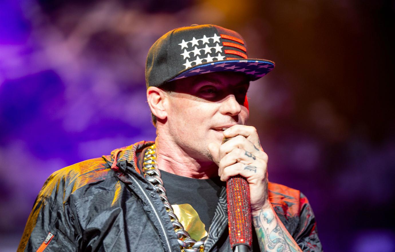 Vanilla Ice on stage holding a mic and wearing a stars and stripes baseball cap, a huge gold chain, and a black jacket.