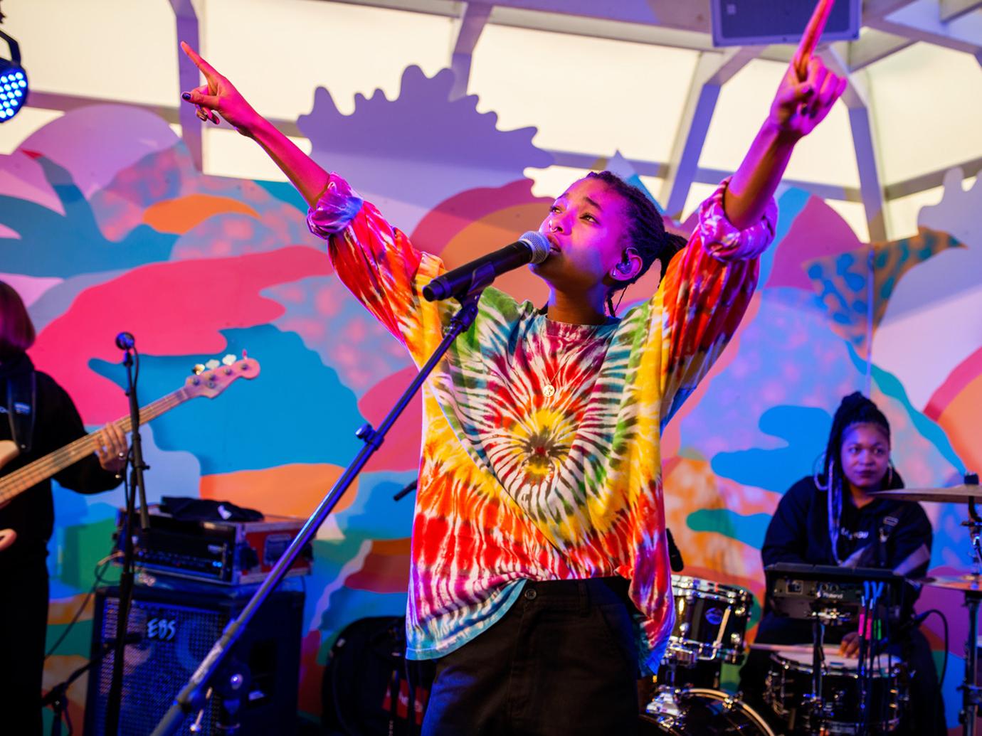 Willow Smith Wearing a Tie-Dye-Shirt Sings On Stage In To Mic With Hands in the Air