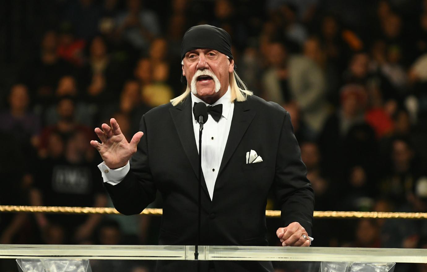 Hulk Hogan stands on stage, in a black tux and black bandanna and talks into a mic.